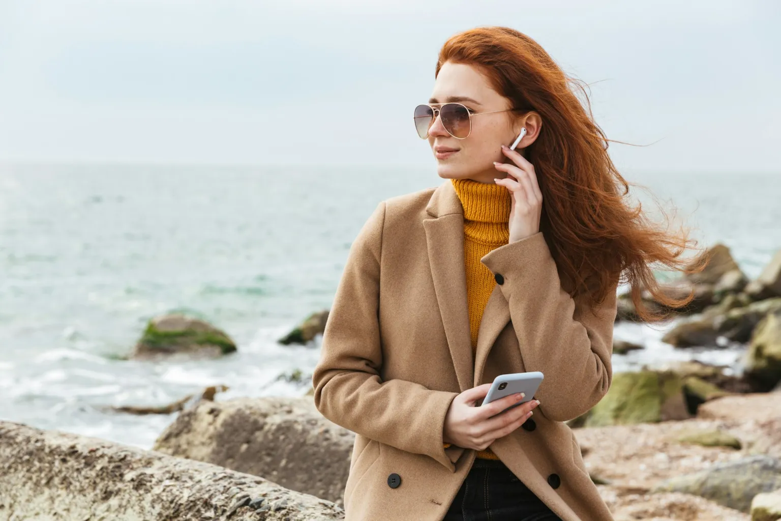 A woman on a rocky seashore listening through wireless earbuds, phone in hand